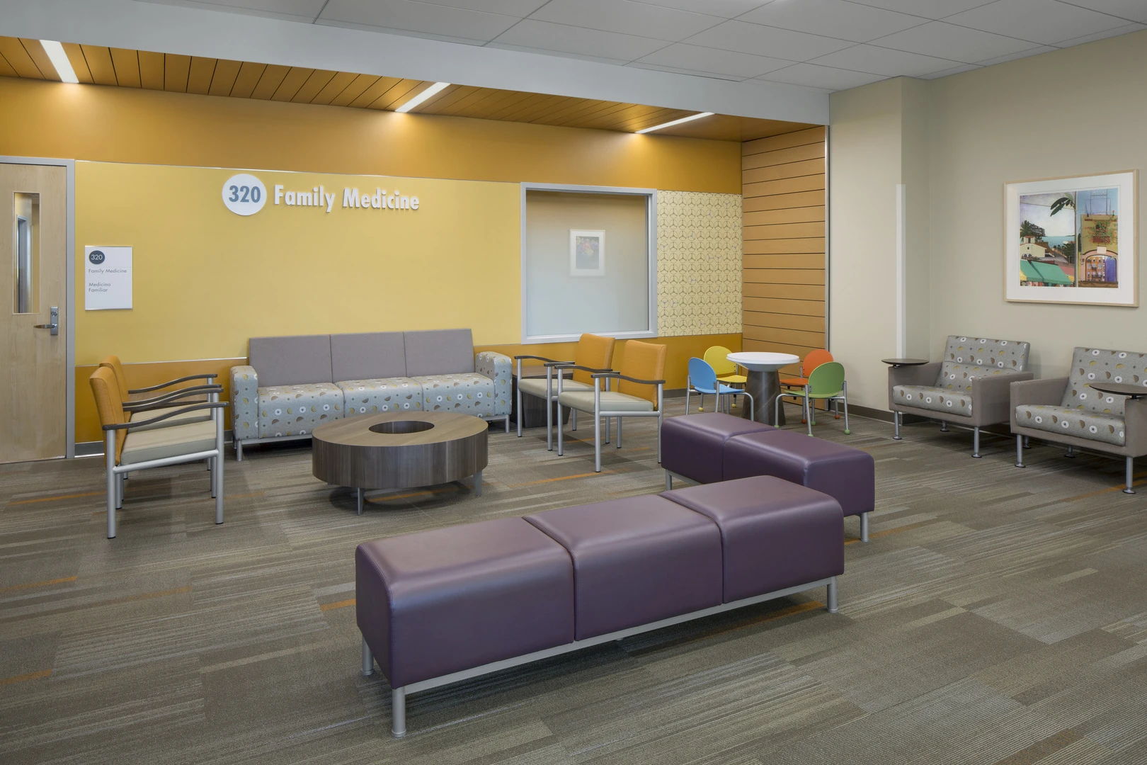 Family Medicine waiting area with a large room number sign, mixed seating including benches, armchairs and sofas, and a small children's table with colorful chairs.