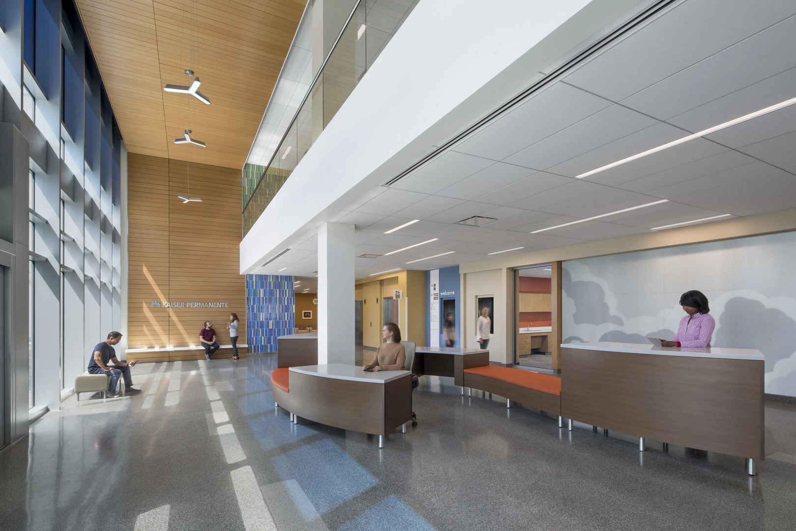 Main lobby at Kaiser Permanente Skyport: a double-height timber-clad wall with Kaiser Permanente signage, curved reception desks with built-in bench seating, and daylight falling across terrazzo flooring.