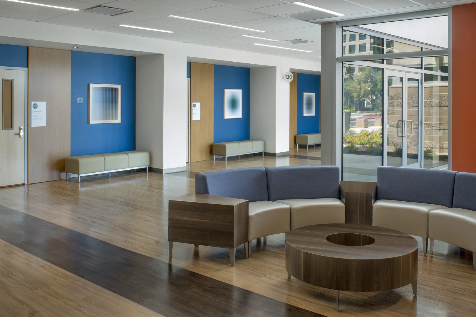 Ground-floor corridor with deep blue accent walls framing conference room doors, built-in benches in each alcove, and curved lounge seating on wood-look flooring.