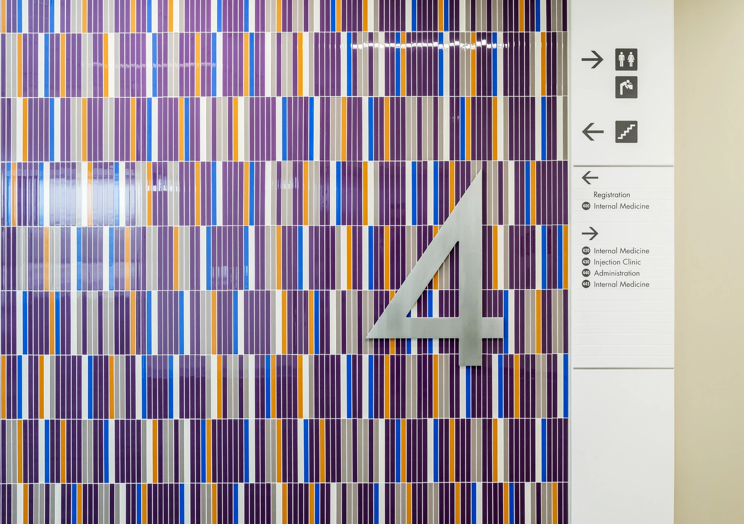 Large brushed-metal number 4 mounted on a striped purple, orange and blue tile wall beside a directional sign listing Registration, Internal Medicine, Injection Clinic and Administration.