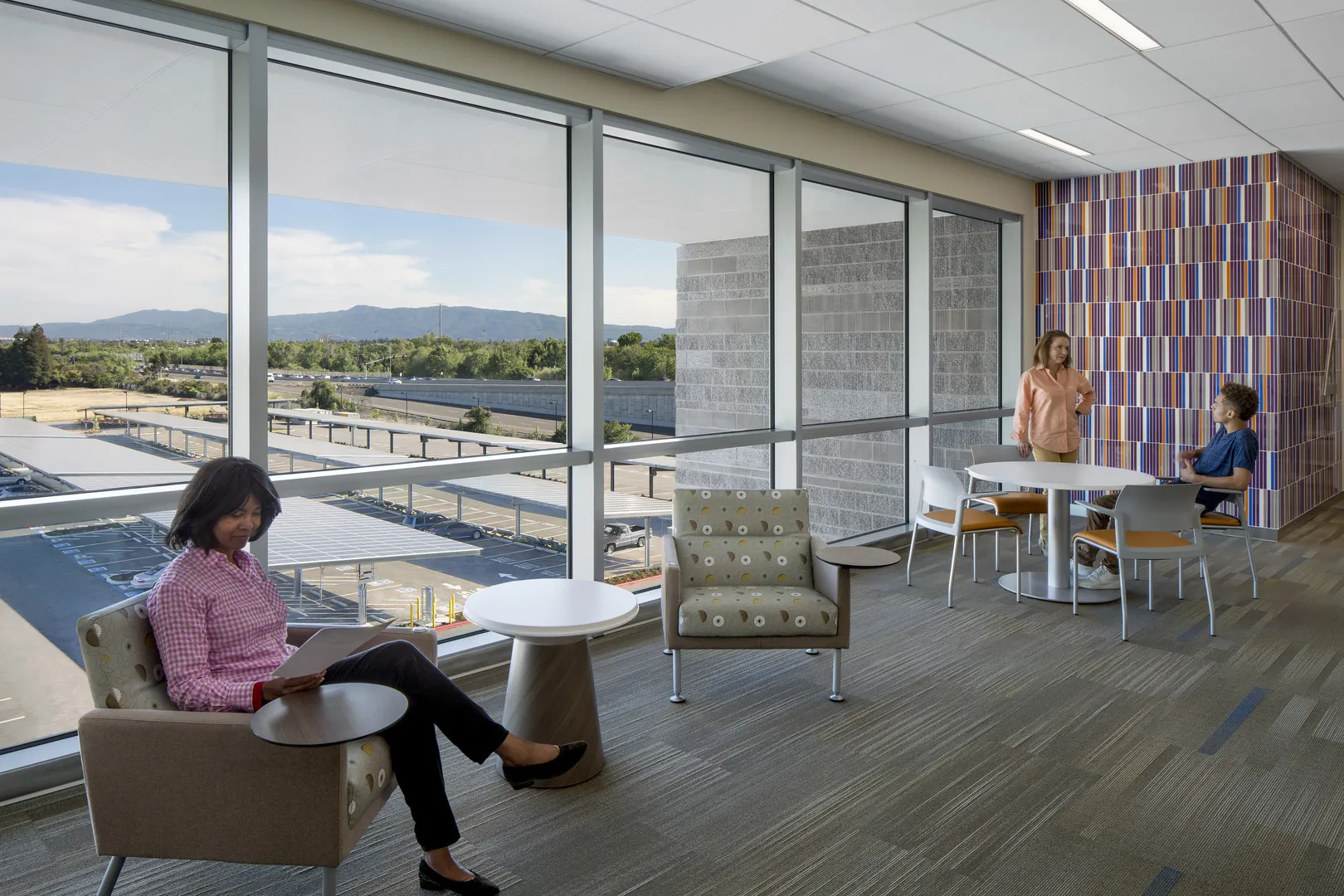Upper-floor lounge with floor-to-ceiling windows overlooking the hills, upholstered lounge chairs, and a small café table beside a striped tile wall.